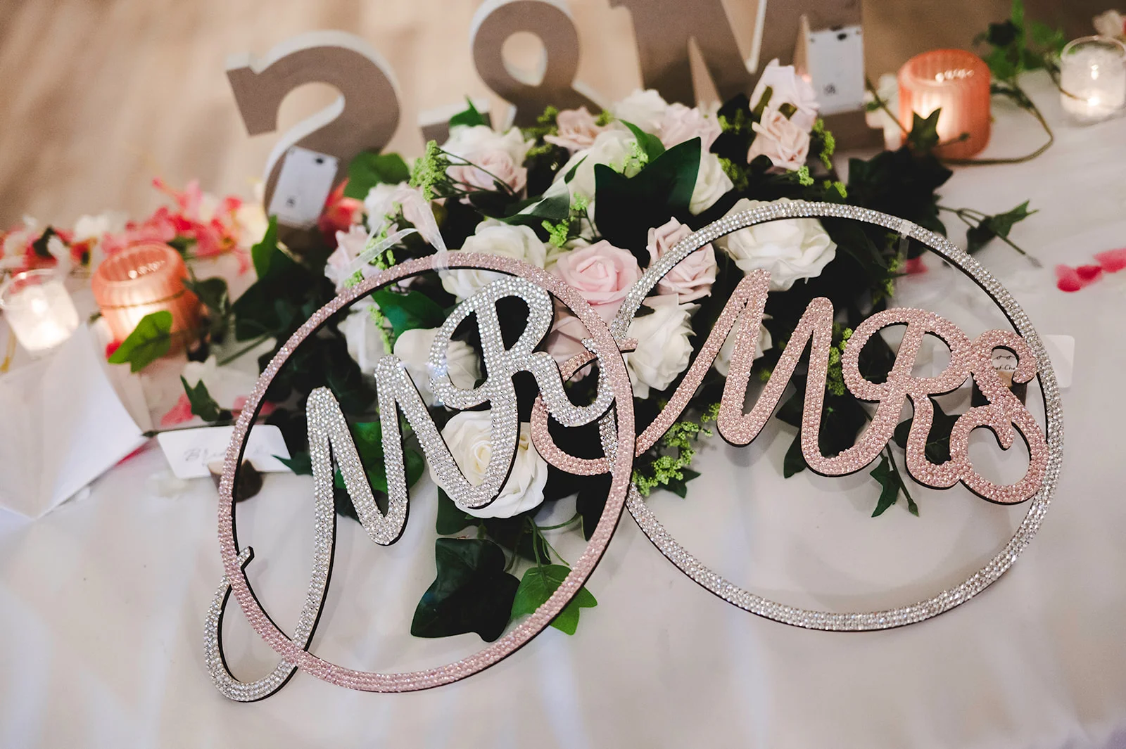 Sparkling crystal mr and mrs hoop signs sit atop a lush white and pink floral arrangement with glowing candles on a wedding reception table.