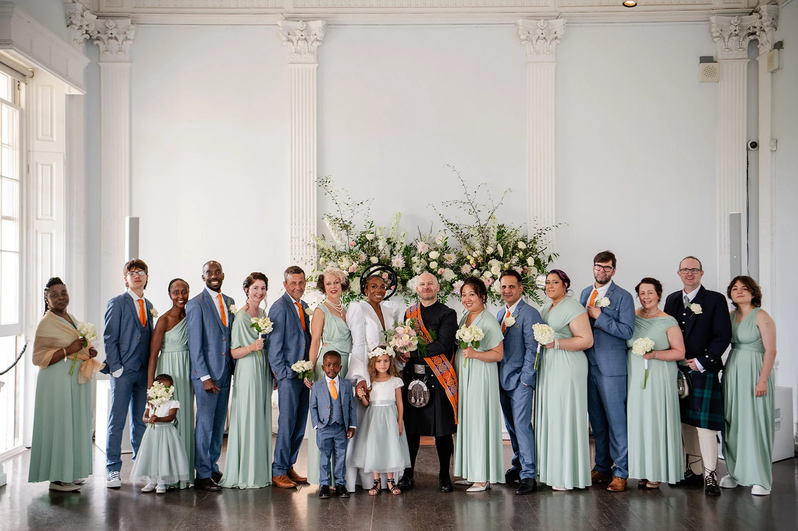 A large wedding party dressed in sage green and blue suits poses formally in front of a grand floral arrangement inside a stately home.