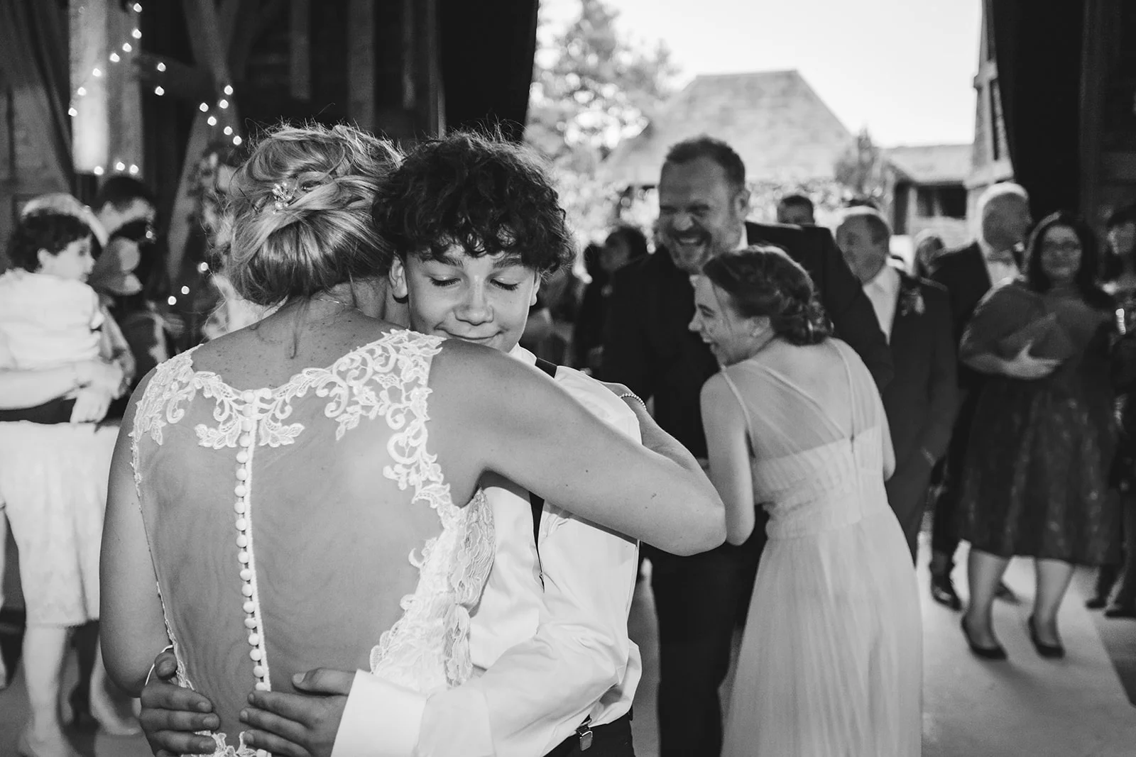 A bride in an illusion-back lace wedding dress shares an emotional embrace and a tender moment with a young guest during a joyful wedding reception.