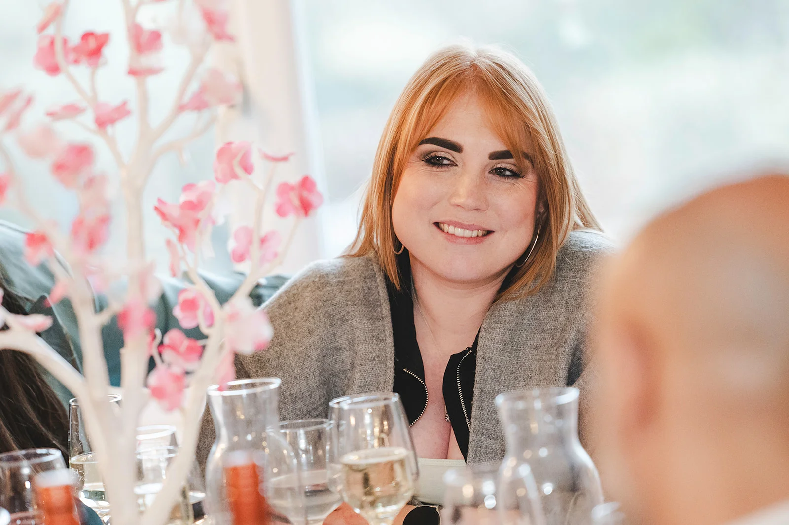 A guest smiles warmly while seated at a reception table adorned with wine glasses and decorative pink blossom branches.