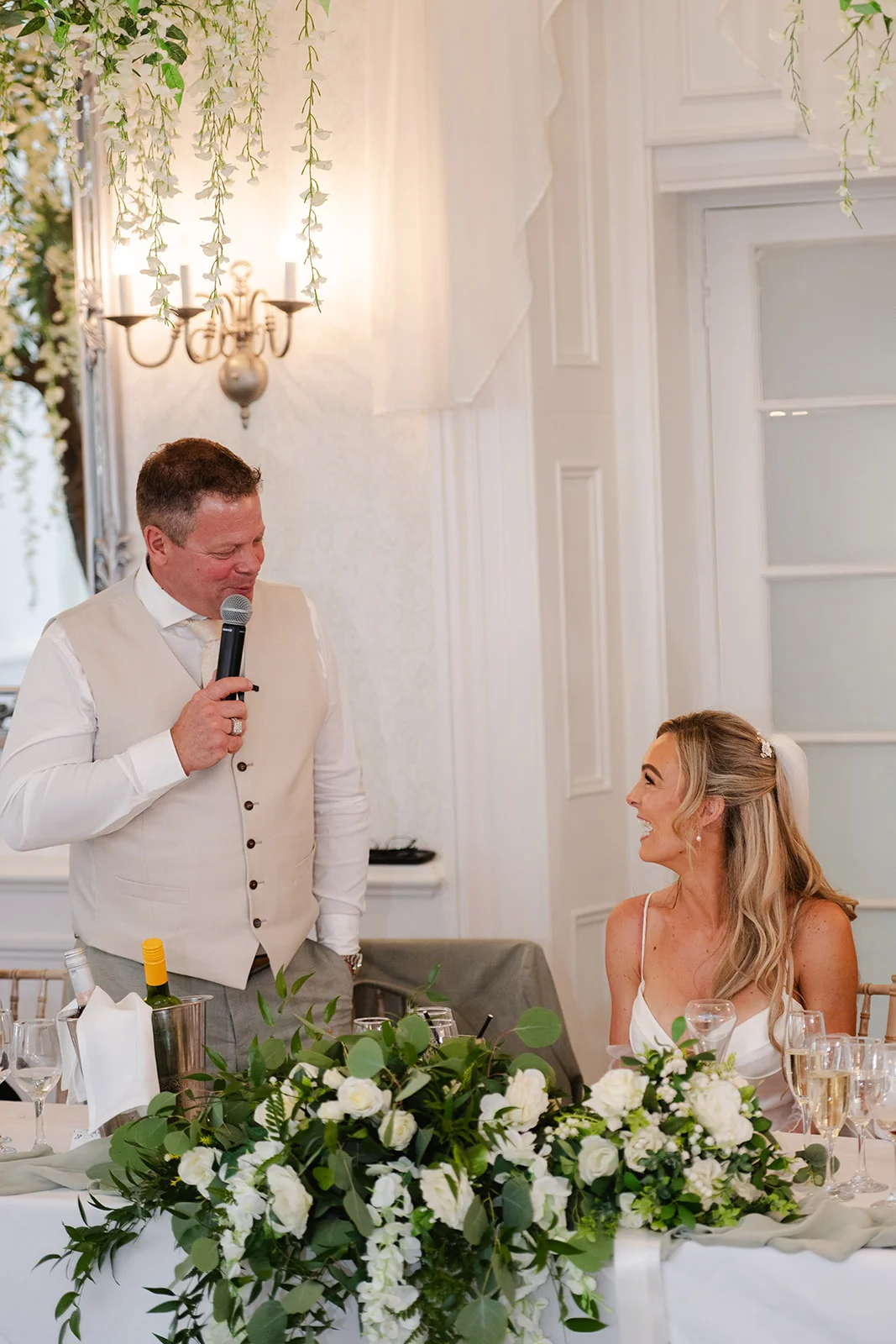 A groom delivers a speech into a microphone at the top table while the laughing bride listens attentively next to a lush white floral arrangement.