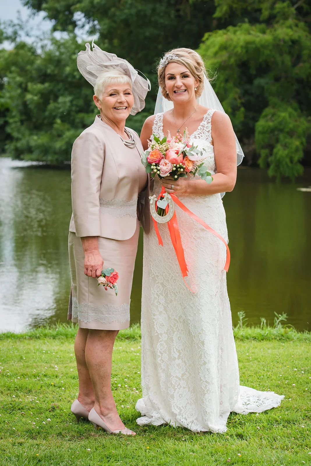 A smiling bride in a lace wedding dress holding a colourful bouquet stands alongside her mother wearing a matching hat and jacket by a lakeside.