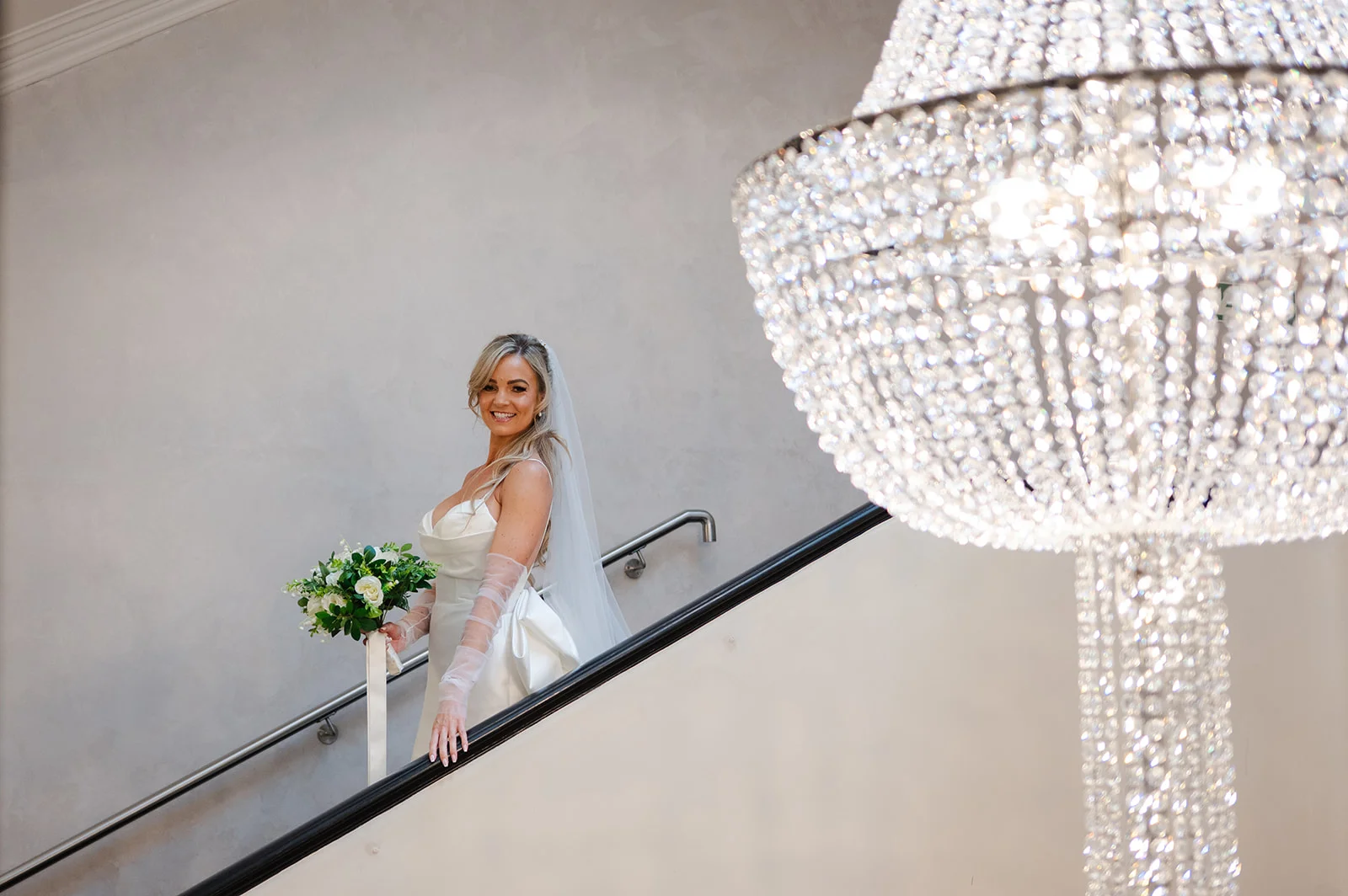 A smiling bride holding a white rose and eucalyptus bouquet stands on a grand staircase next to a large sparkling crystal chandelier.