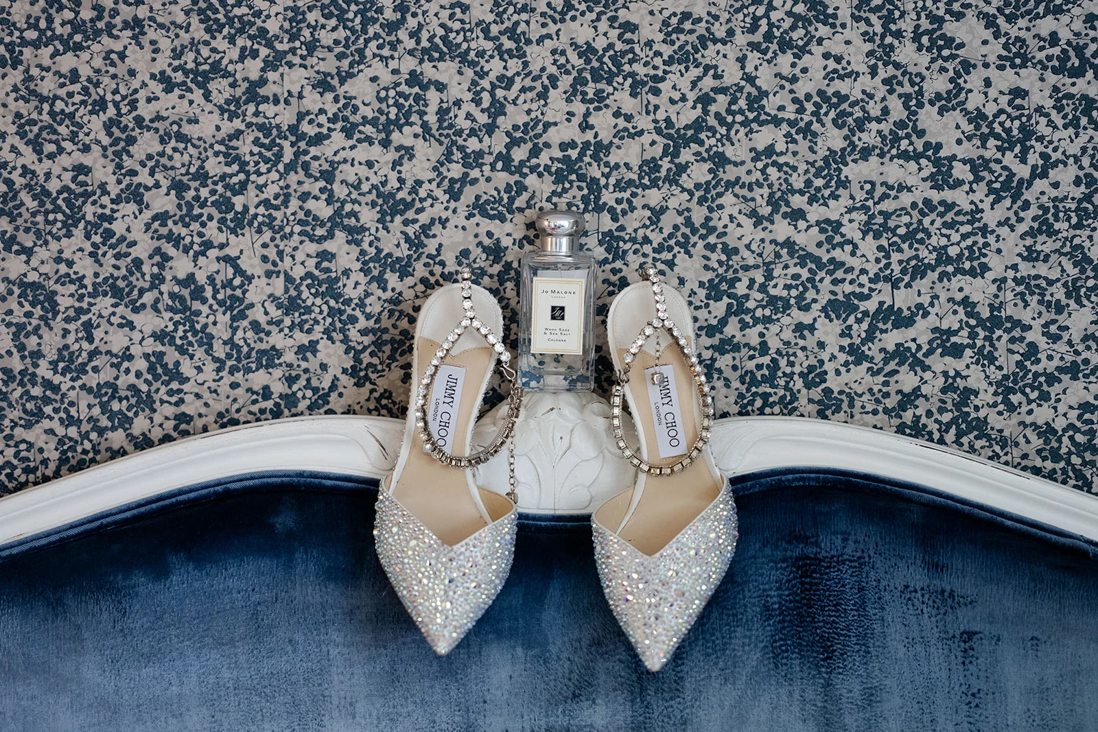 A bridal flat lay featuring crystal-embellished pointed heels resting on a blue velvet surface against a patterned wallpaper backdrop alongside a perfume bottle.