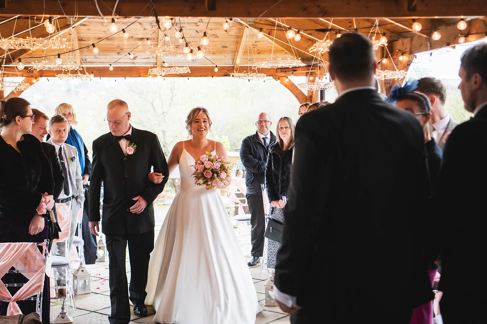 A smiling bride walks down the aisle on her father's arm beneath a wooden pavilion strung with fairy lights towards the waiting groom.