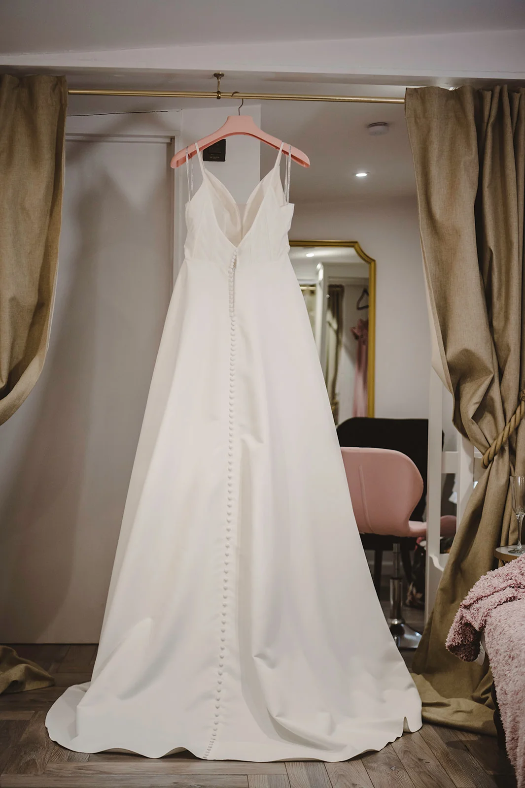 An elegant satin a line wedding dress featuring spaghetti straps and a long row of delicate buttons down the back hangs framed by curtains in a bridal dressing room.