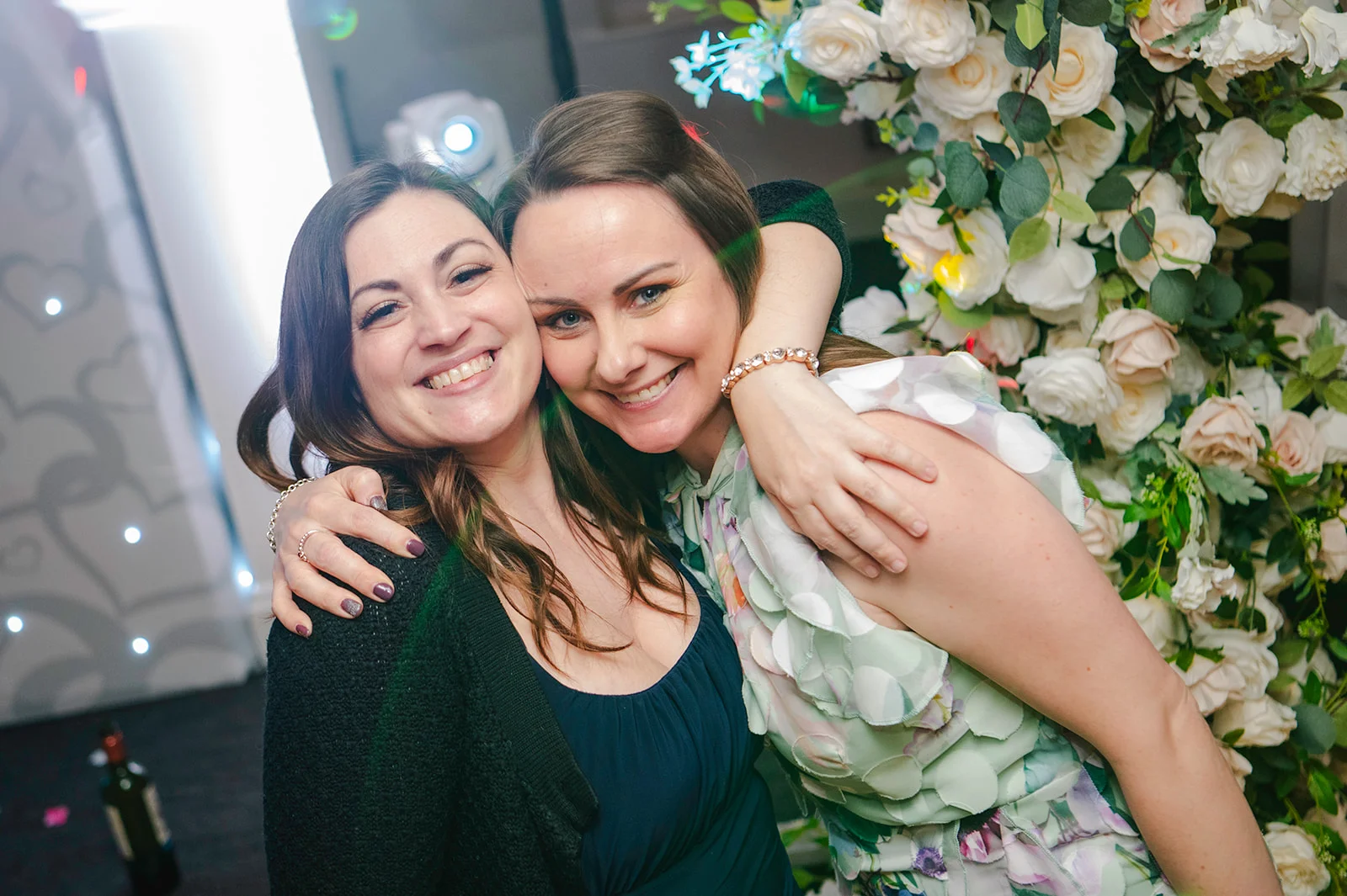 Two smiling female wedding guests embrace warmly in front of a lush white rose and floral wall installation during the evening celebration.
