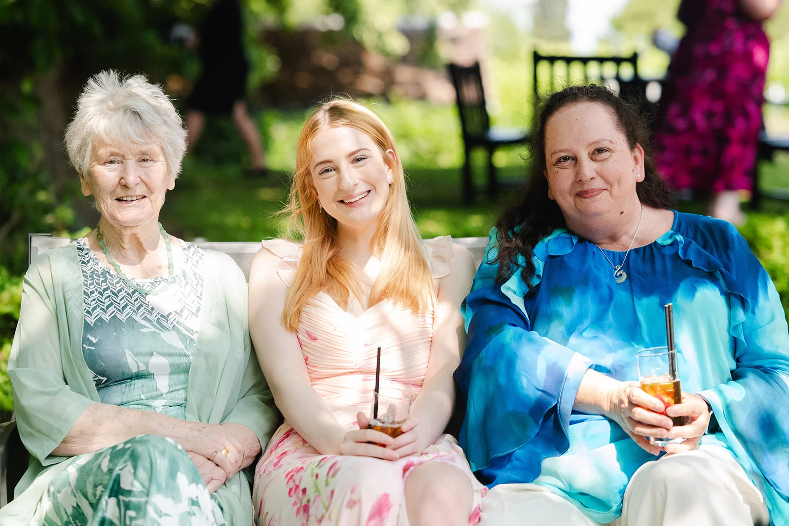Three female wedding guests sit together on an outdoor bench in dappled sunlight, smiling and holding drinks during the reception.