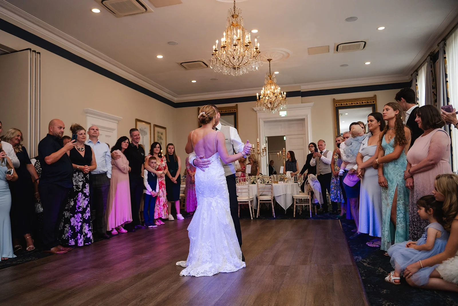 The newlywed couple share their first dance on a wooden dance floor in a ballroom surrounded by watching guests.
