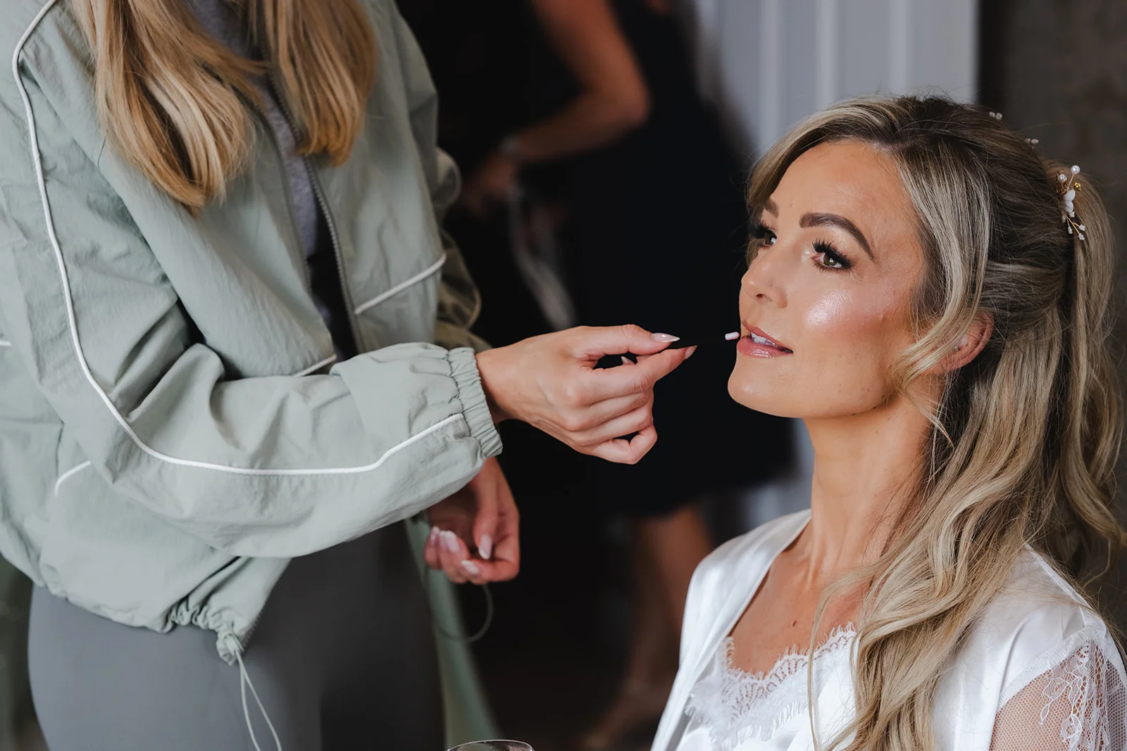 A professional makeup artist carefully applies lipstick to a bride during morning bridal preparations as she wears a white lace-trimmed robe.