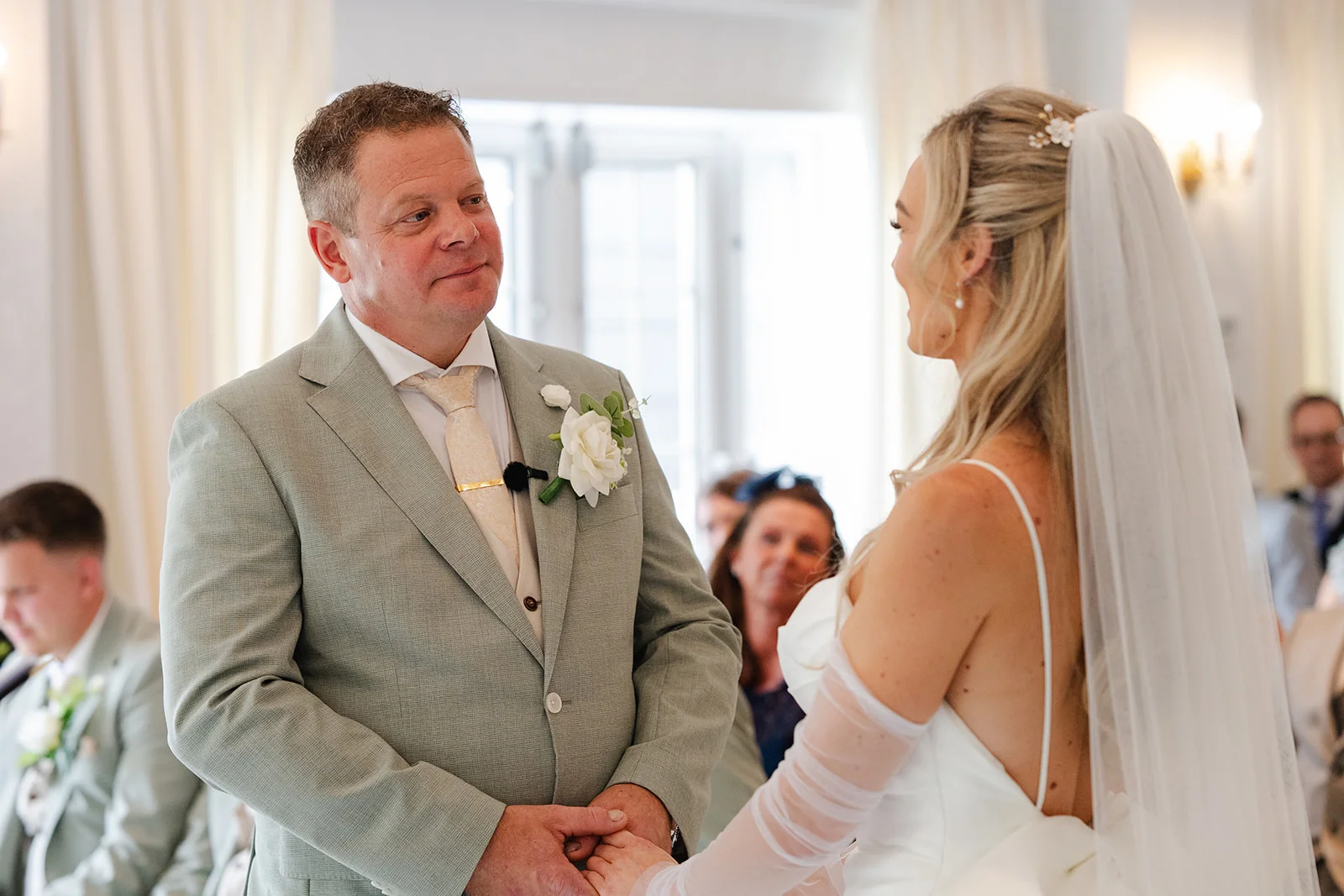 An emotional groom in a sage suit looks lovingly at his bride holding hands during their wedding ceremony.