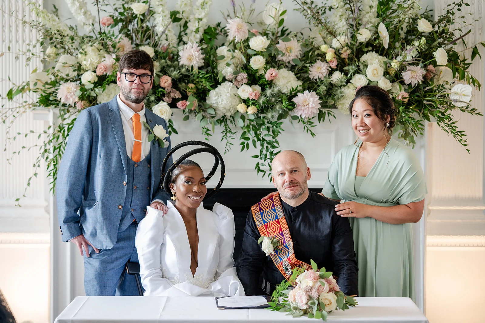 A smiling interracial couple and their wedding party pose together behind a signing table beneath a grand floral mantelpiece installation.