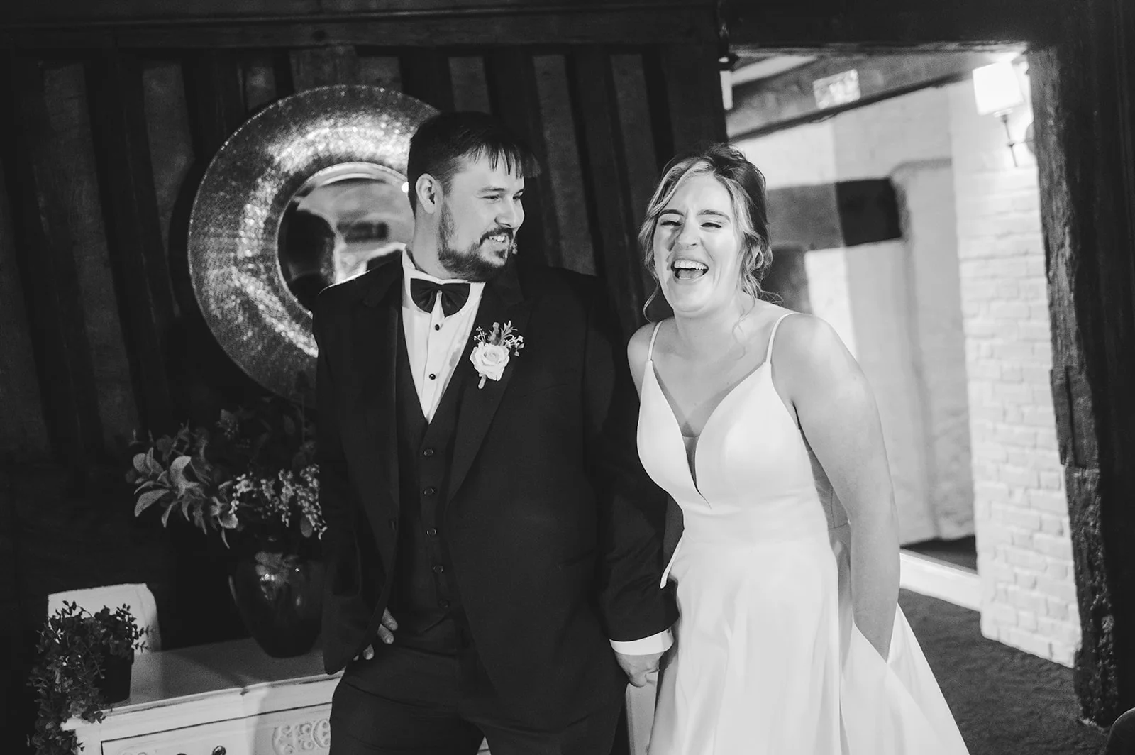 A newlywed couple laugh joyfully as they walk hand in hand through an atmospheric barn venue captured in a candid black and white shot.