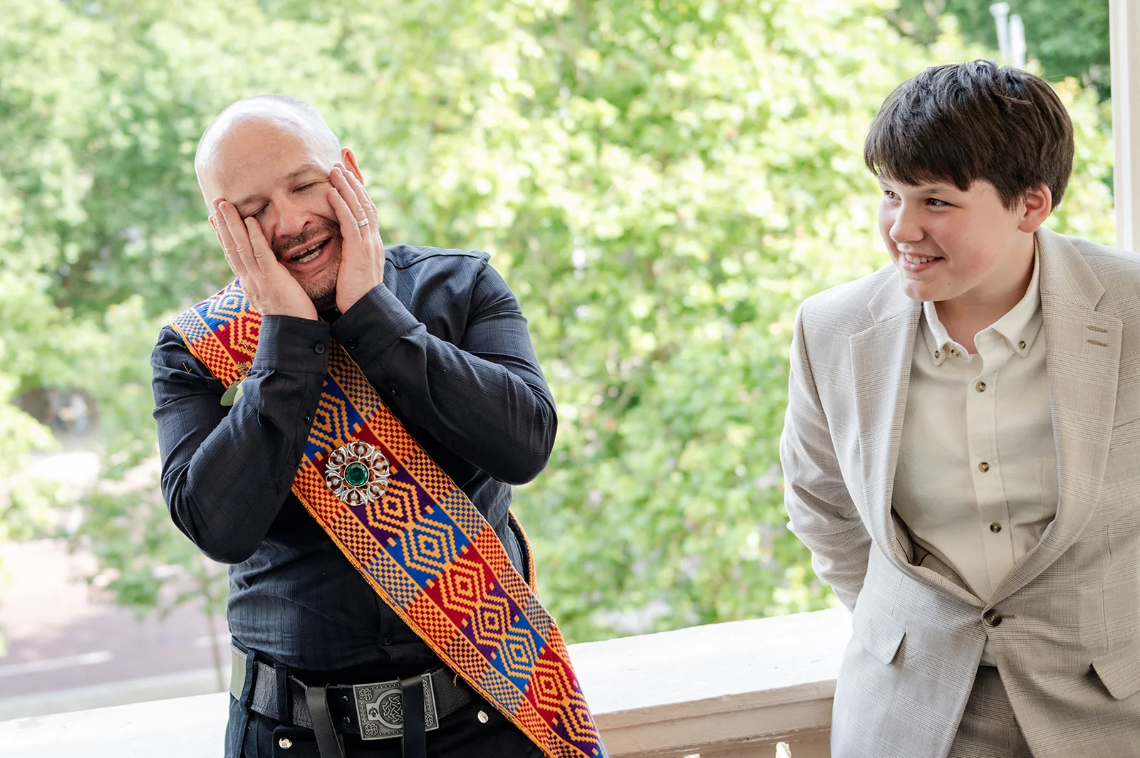 An emotional man laughs with his hands on his face while wearing a patterned tartan sash, standing next to a smiling young person in a light grey suit.
