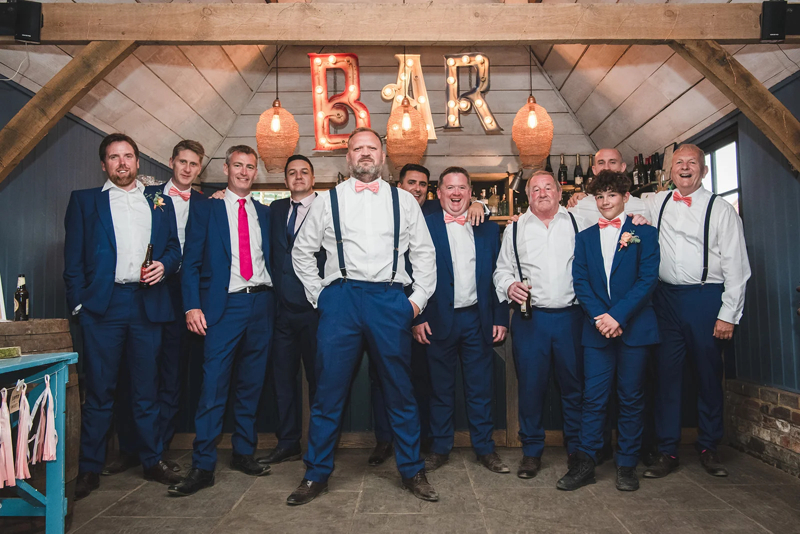 A group of groomsmen in blue suits and matching pink bow ties pose happily together in front of a rustic wooden bar illuminated by a lighted bar sign.