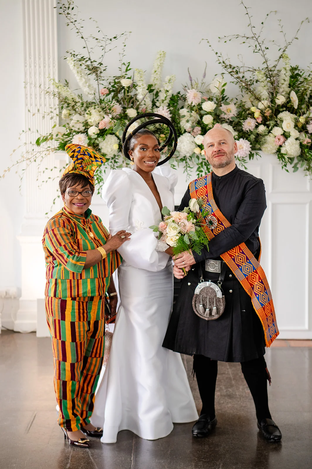 A newlywed couple posing with a guest against a large floral display indoors, featuring a bride in a white gown and sculptural halo headpiece, a groom in a black kilt with a patterned tartan sash, and a guest in vibrant striped cultural attire.