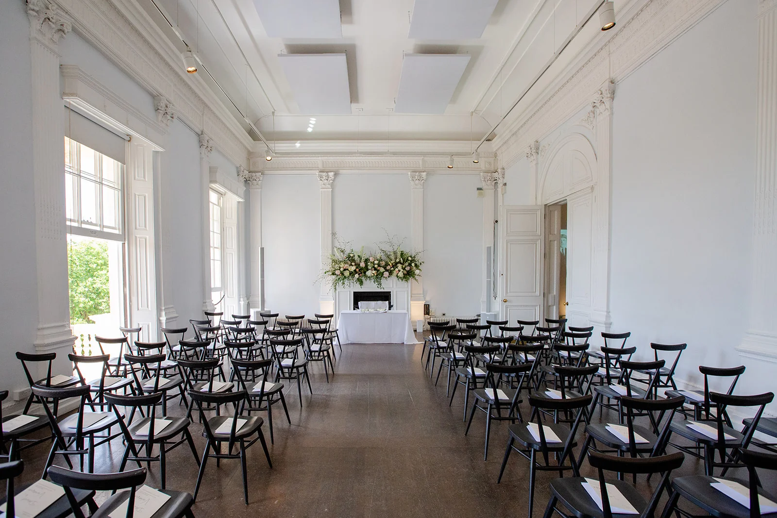 An elegant civil ceremony venue features high ceilings, classic white walls, and rows of black chairs arranged facing a decorated fireplace mantelpiece.
