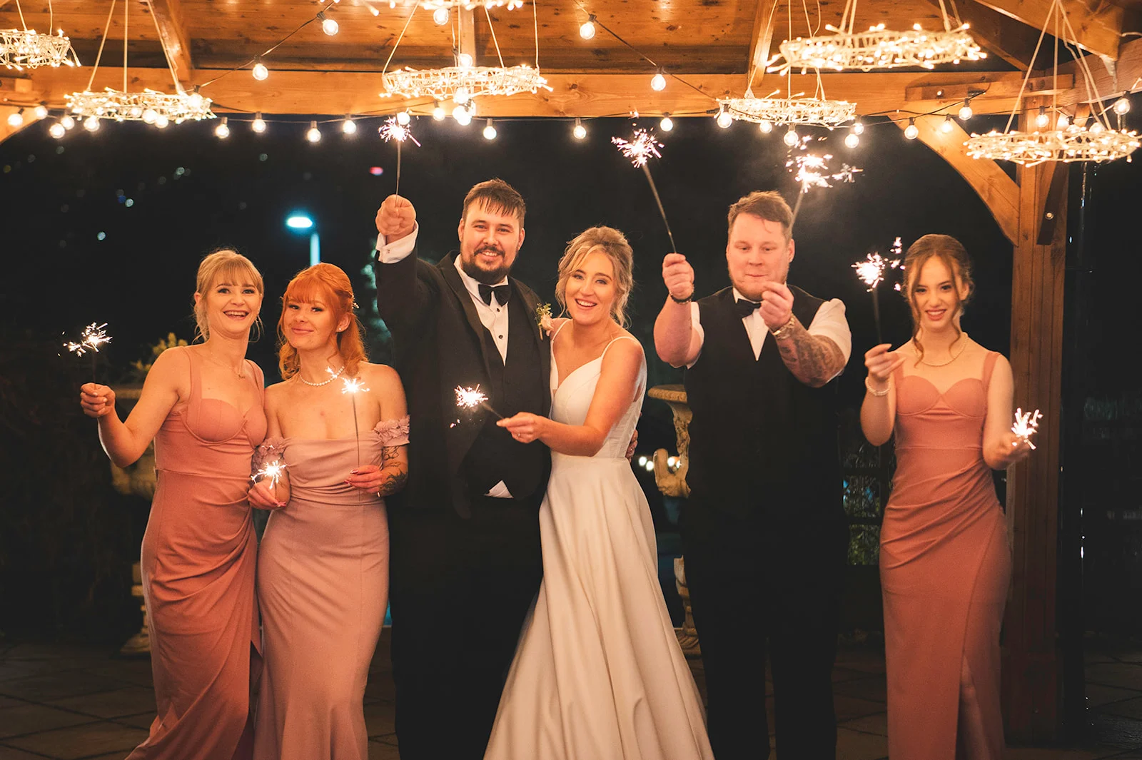 A newlywed couple and their wedding party pose together at night under a wooden structure lit by fairy lights, holding sparklers.