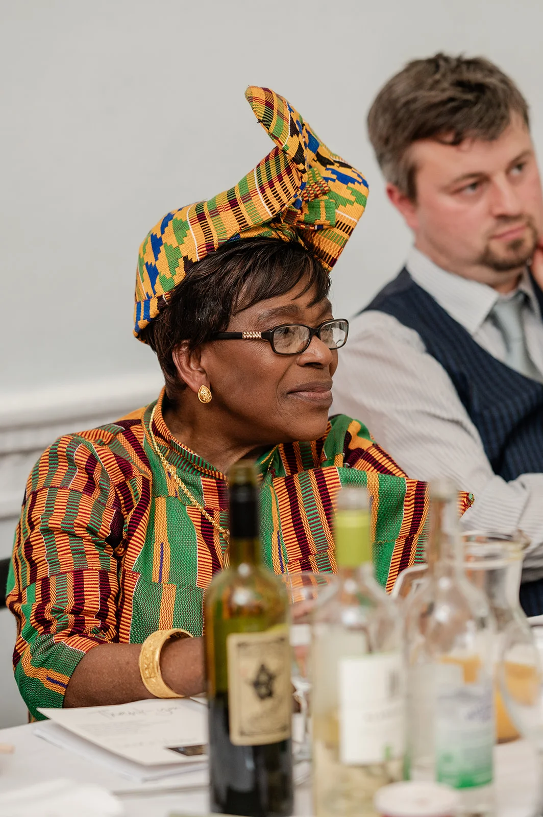 An elegantly dressed guest wearing a colourful kente cloth outfit and matching headwrap sits smiling during the wedding reception.