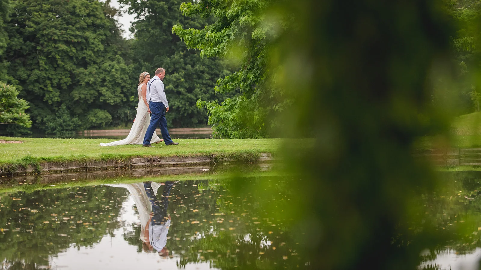 A newlywed couple walks hand in hand along a grassy bank beside a calm lake, their reflections visible in the water, framed by foliage in the foreground.