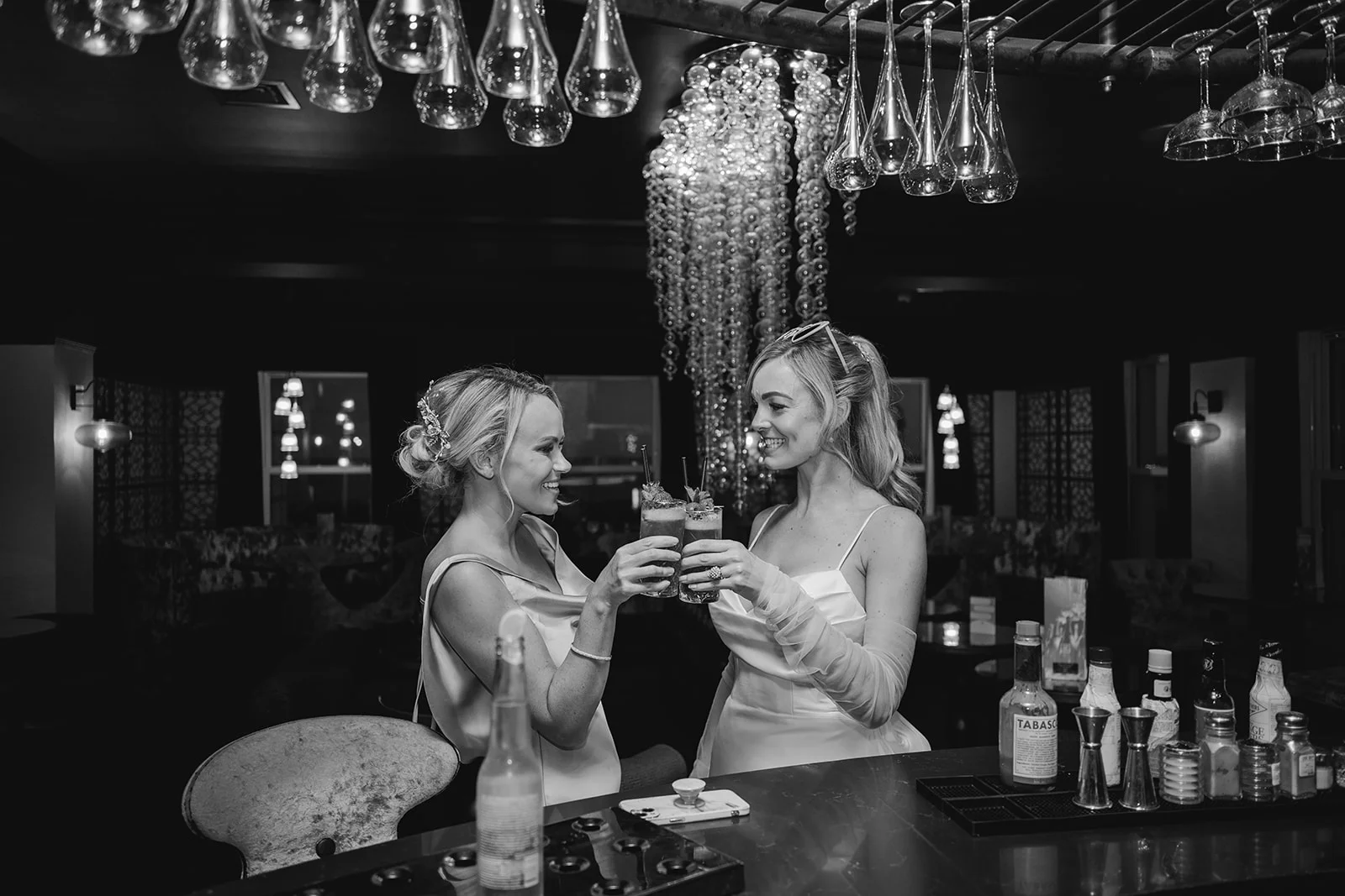 Two smiling brides clink cocktail glasses across a stylish bar counter during their wedding reception celebration.