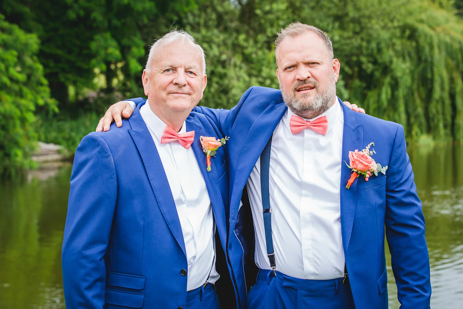 An older same-sex couple wearing matching blue suits and coral bow ties pose affectionately with their arms around each other by a tranquil lakeside.
