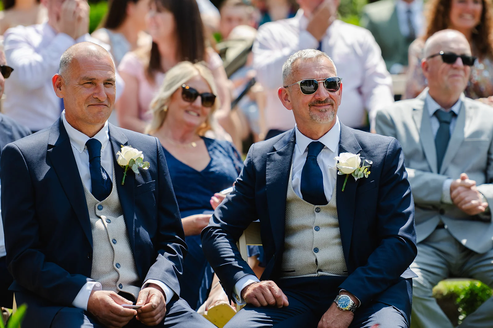 Two smiling grooms in matching navy suits and waistcoats sit among wedding guests during an outdoor ceremony.