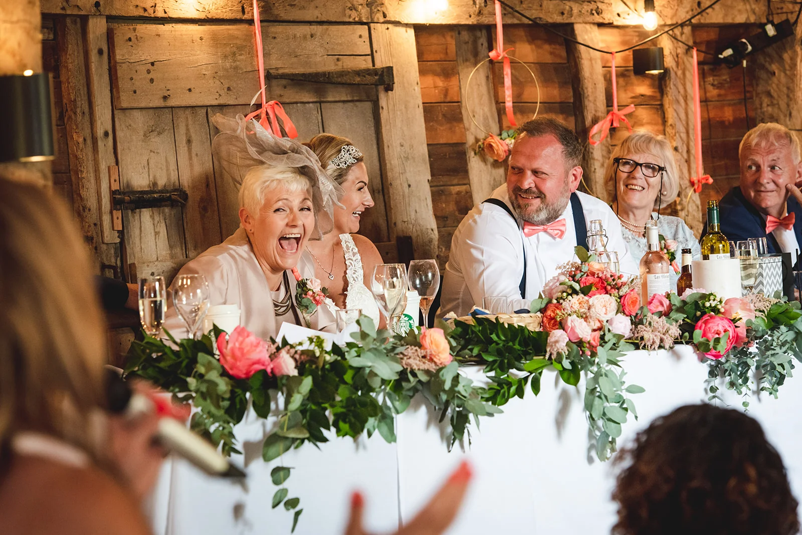 The bride and groom seated at a top table in a rustic barn venue share a joyful laugh during the wedding speeches, surrounded by family and floral decorations.