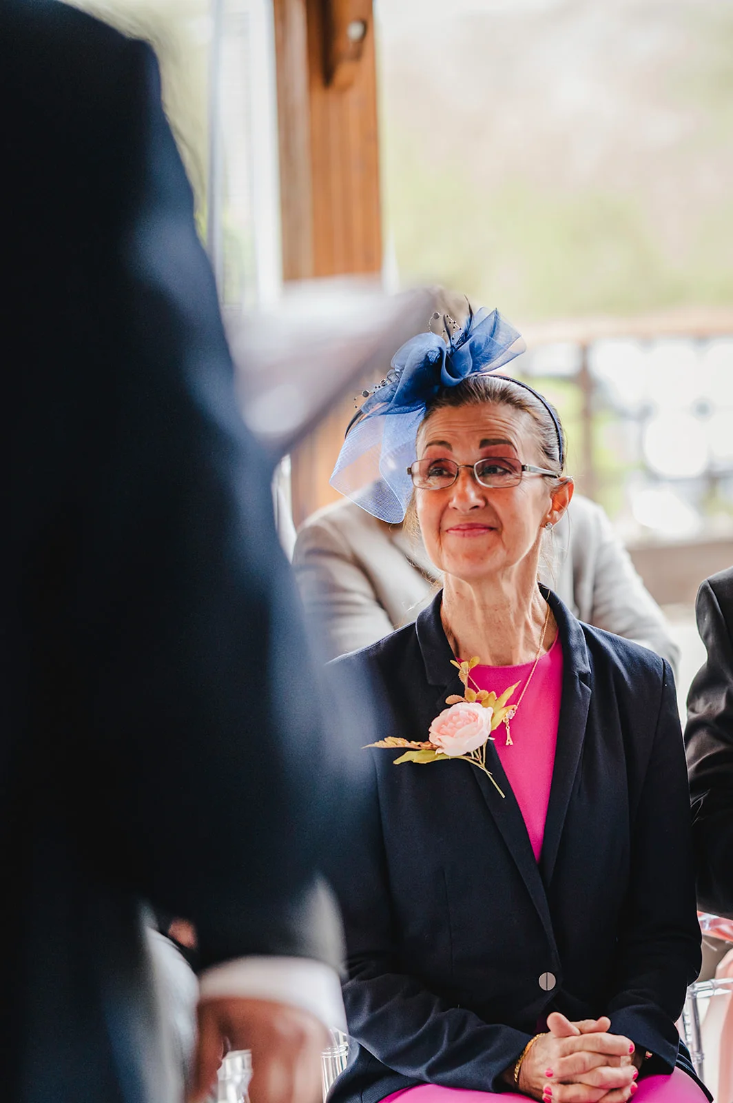 An older woman wearing a navy jacket, pink top, and a blue fascinator watches attentively during a wedding ceremony, holding her hands together.