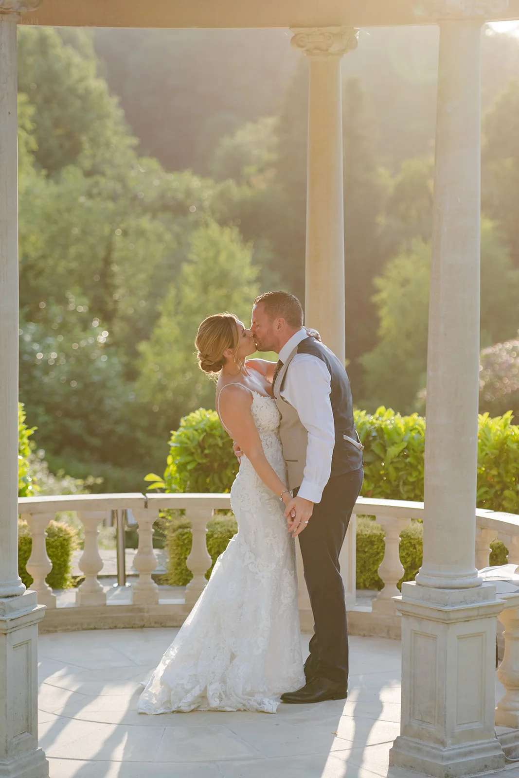 Newlyweds share a romantic kiss inside a classical stone gazebo bathed in warm golden hour sunlight.