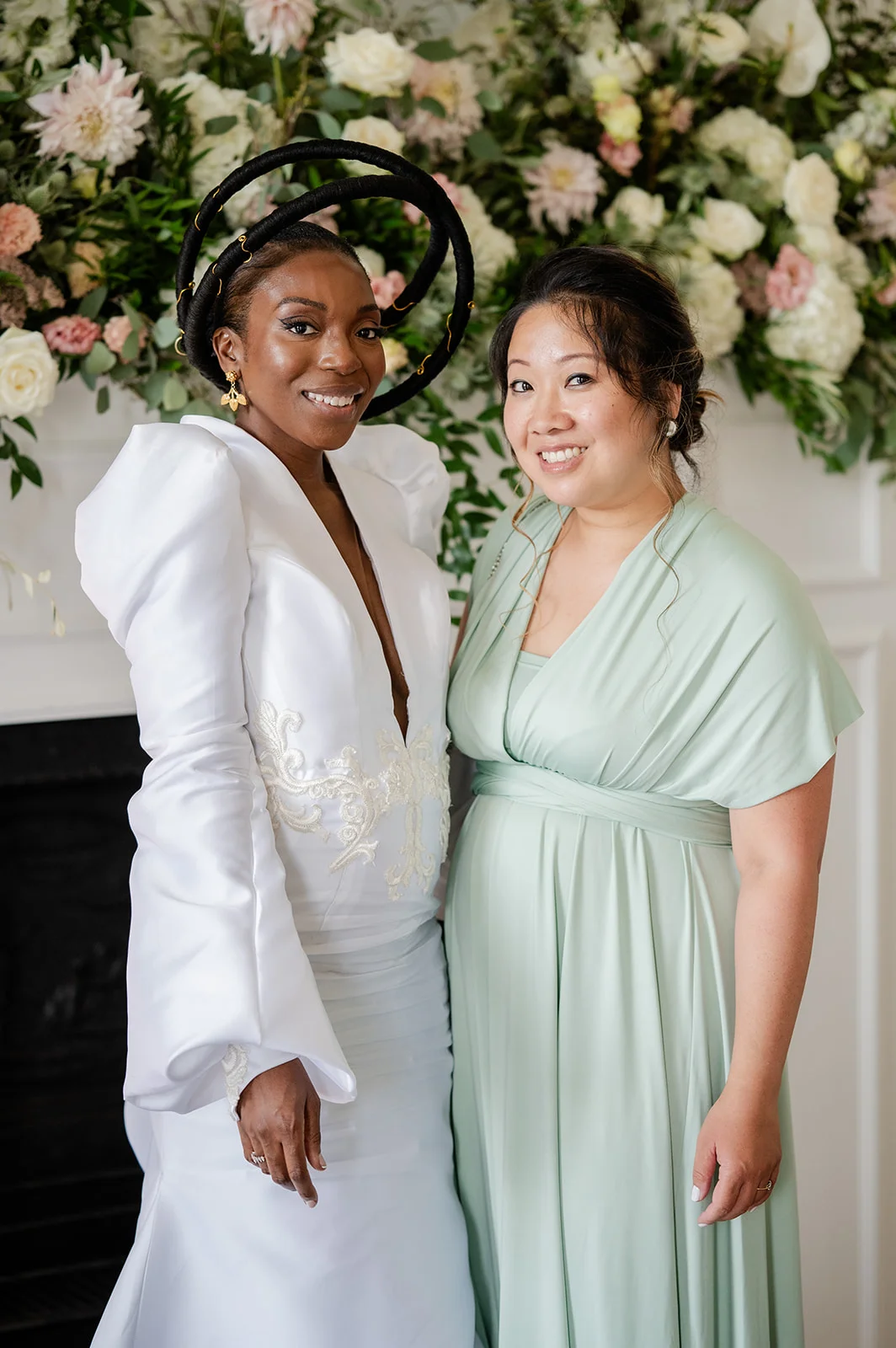 Two smiling brides pose together in front of a lush floral mantelpiece arrangement, one wearing an avant-garde white gown with statement sleeves and sculptural headwear while the other wears a flowing sage green dress.