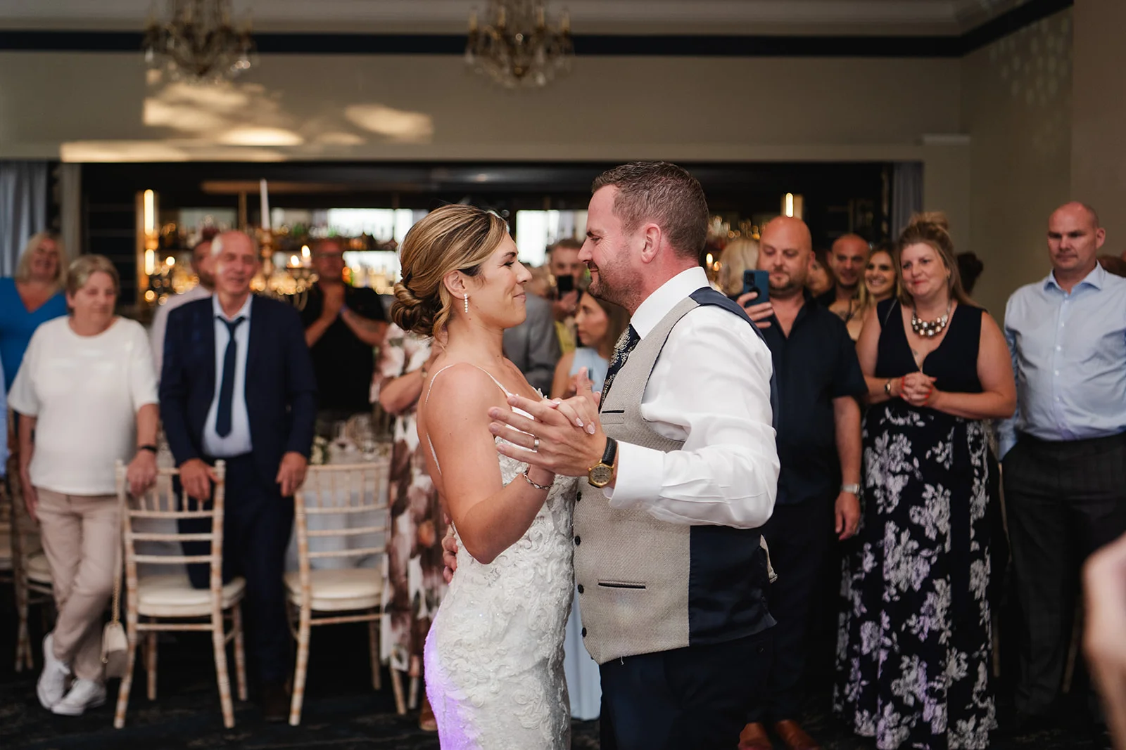 Newlyweds share their first dance together on the dance floor while wedding guests stand around watching and smiling.