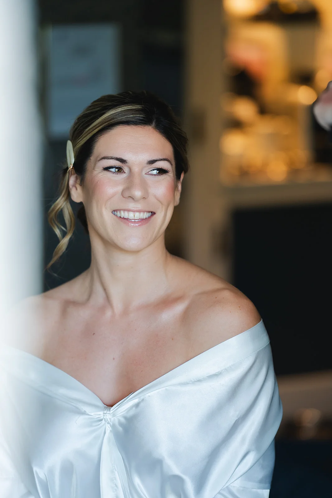 A smiling bride wearing a satin getting-ready robe looks away from the camera in natural light during her morning preparations.