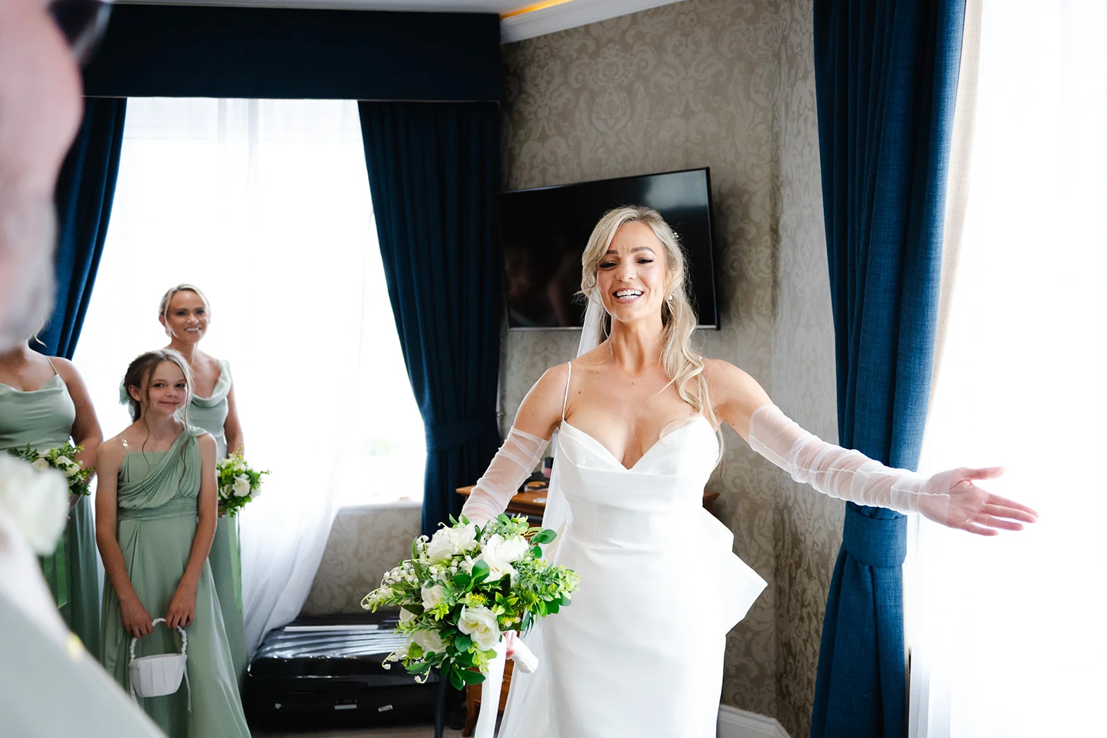 A joyful bride laughs and holds out her arms in a hotel room while showing her white gown and bouquet to her smiling bridesmaids and flower girl.