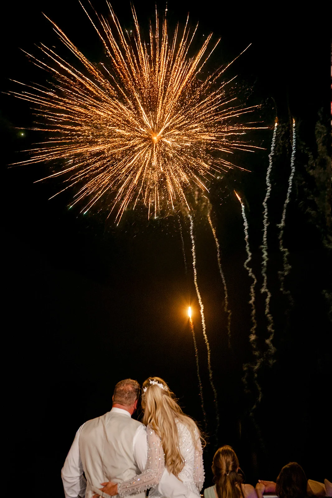 A newlywed couple viewed from behind stands with their arms around each other, watching a spectacular golden firework burst across the night sky.