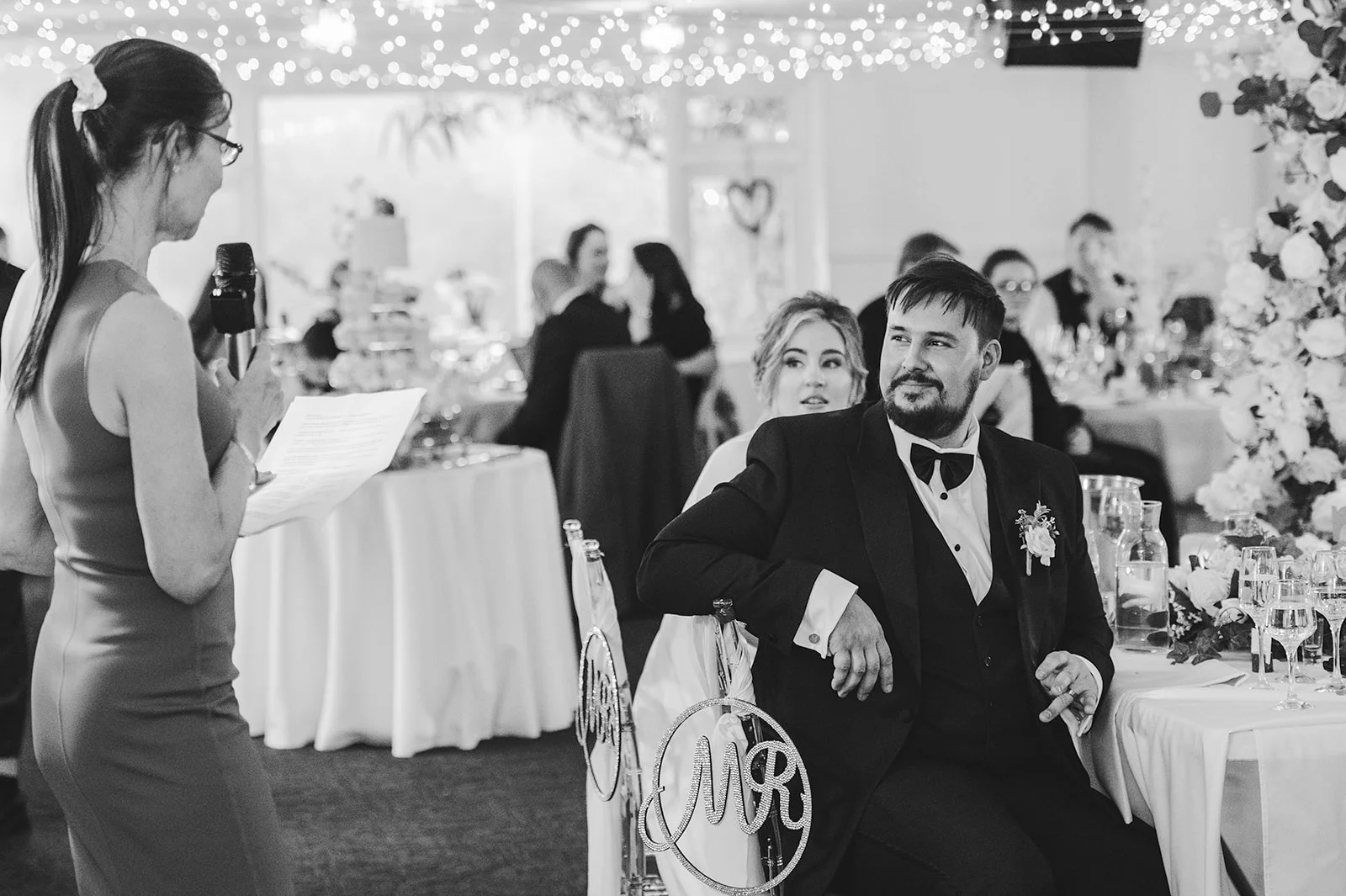 A groom in a black tuxedo sits at the top table listening to a speech while guests dine in a beautifully decorated reception room.