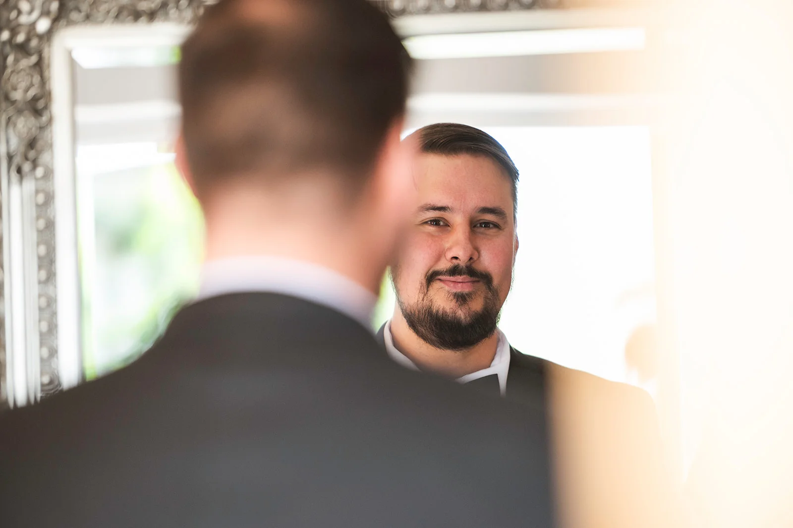 A groom in a black tuxedo and bow tie smiles as he looks into a mirror during his morning preparations.
