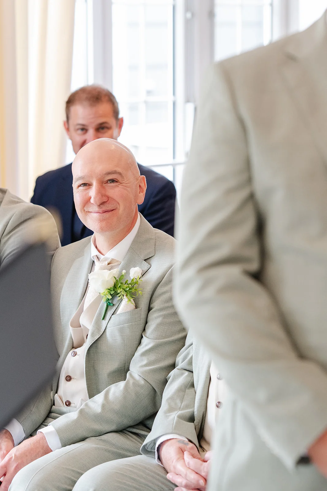 A smiling groom in a sage suit with a white rose buttonhole looks forward during a light and airy indoor wedding ceremony.