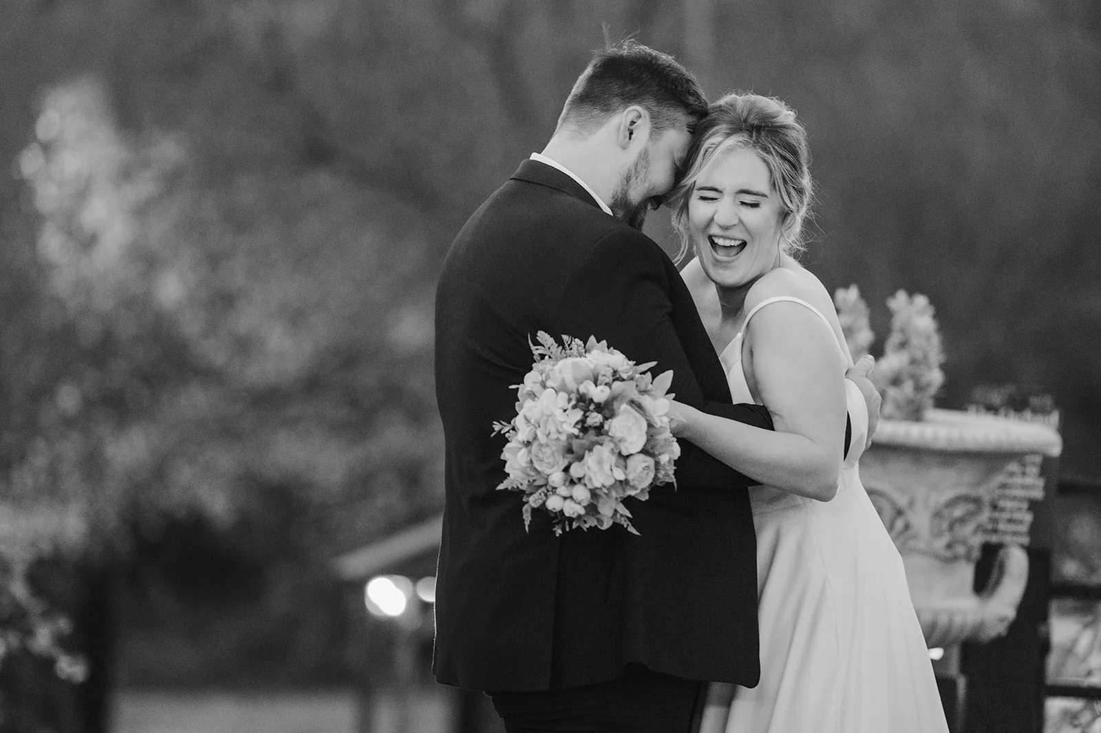 A joyful bride and groom share a tender embrace and a big laugh outdoors while holding a beautiful floral bouquet.