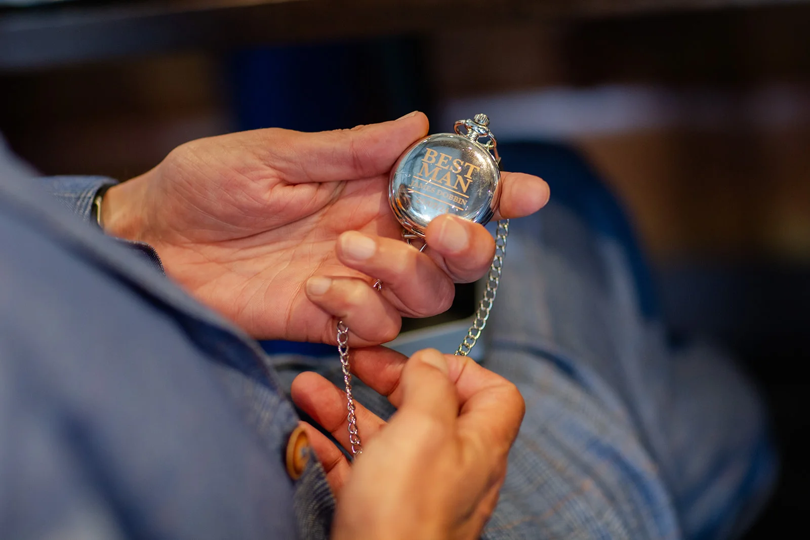 A close-up shot of hands holding a polished silver pocket watch engraved with the words best man and a chain.