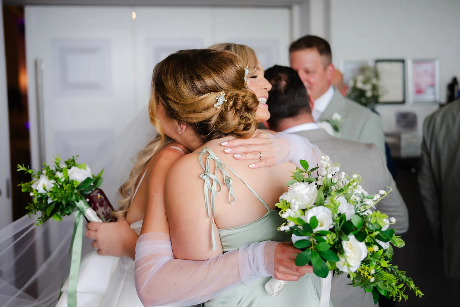 A joyful bride shares a warm, emotional embrace with her bridesmaid holding matching bouquets of white roses and green foliage.