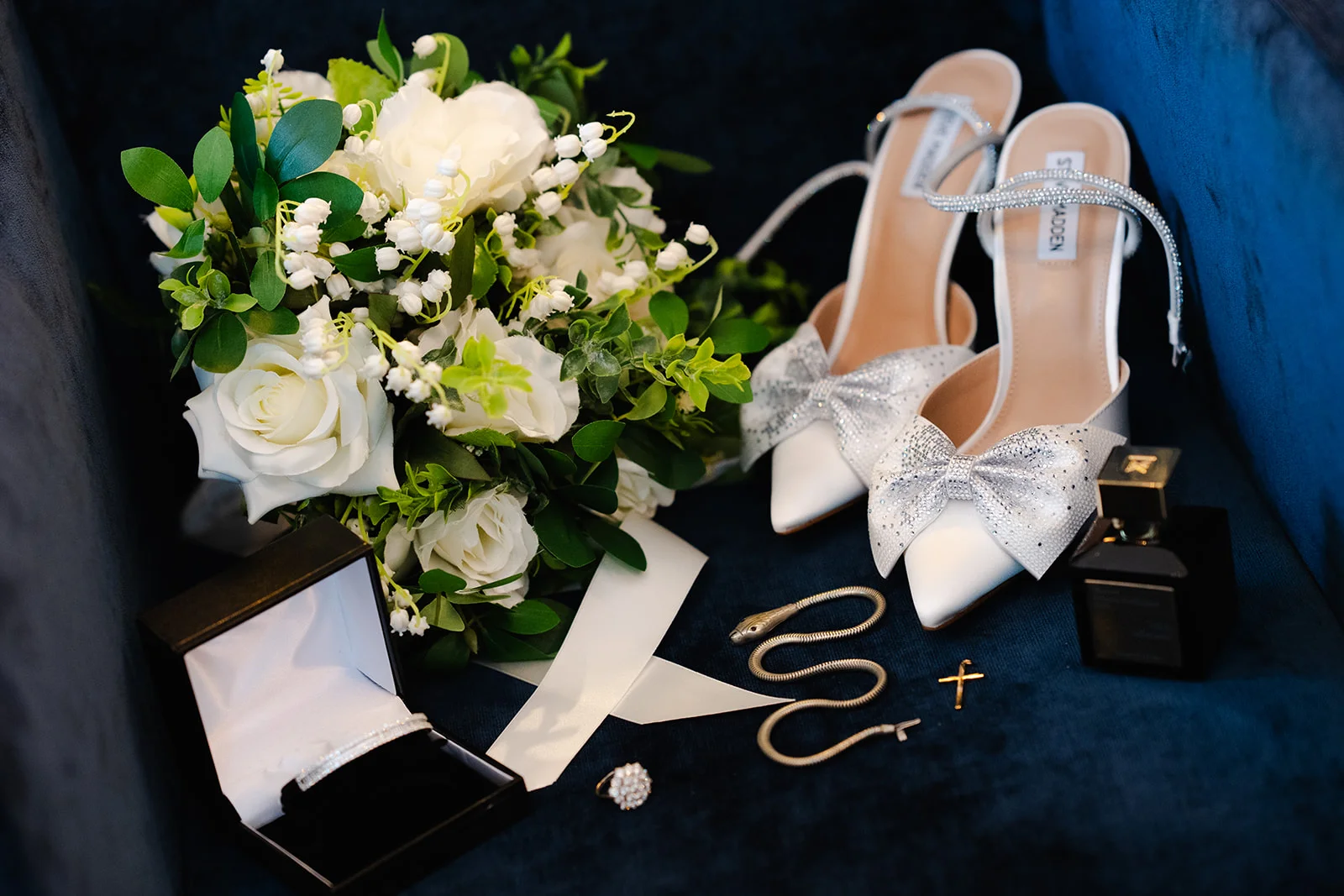 An elegant bridal flat lay features a bouquet of white roses and lily of the valley alongside sparkling bow-embellished heels, a ring box, perfume and jewellery on a dark velvet surface.