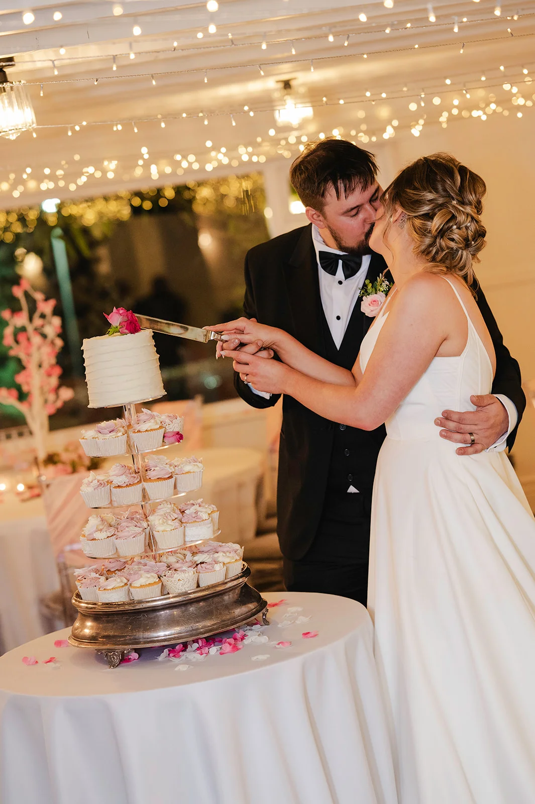 A newlywed couple share a tender kiss while cutting a tiered wedding cake adorned with cupcakes during their evening reception.