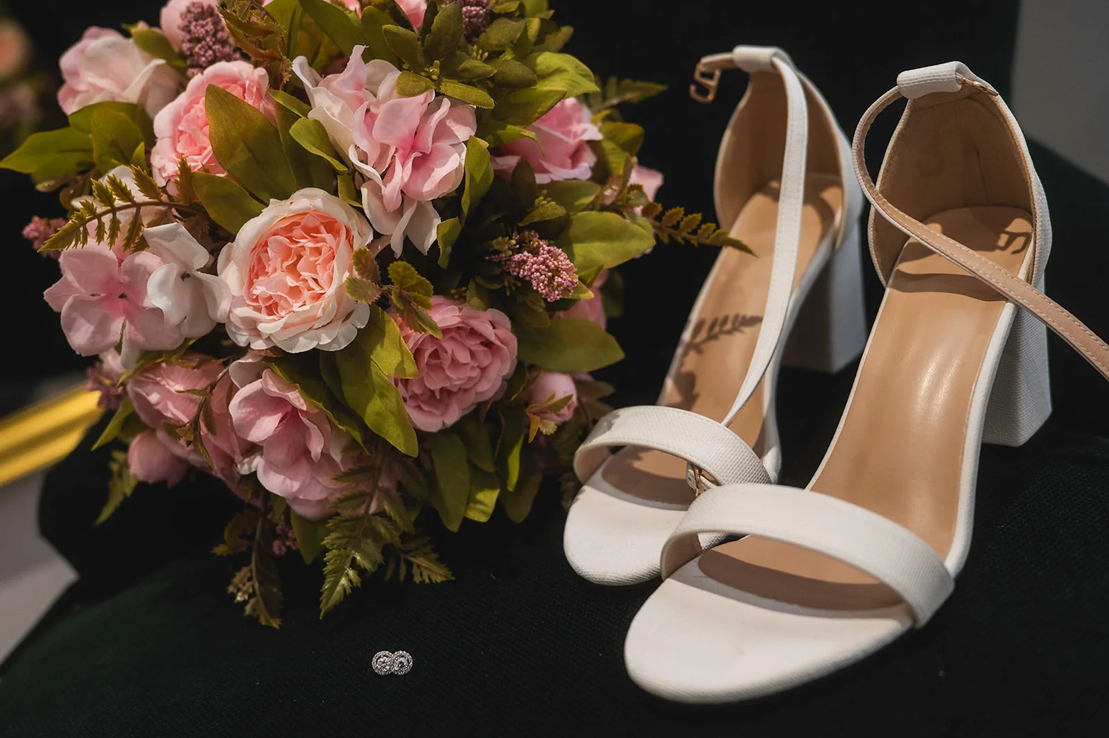 A bridal flat lay featuring a pink floral bouquet next to white block heel sandals and small diamond earrings on a dark surface.