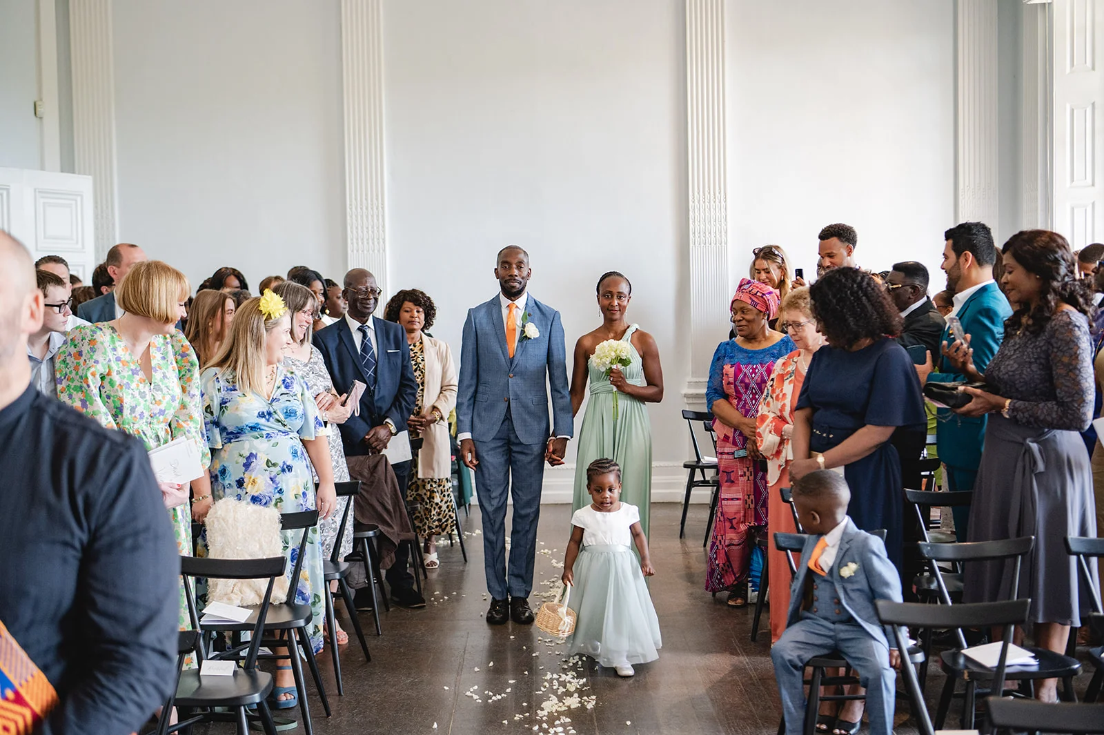 A groom in a blue suit walks down the aisle hand-in-hand with a bridesmaid in a sage green dress, followed by a flower girl scattering petals amongst seated wedding guests.