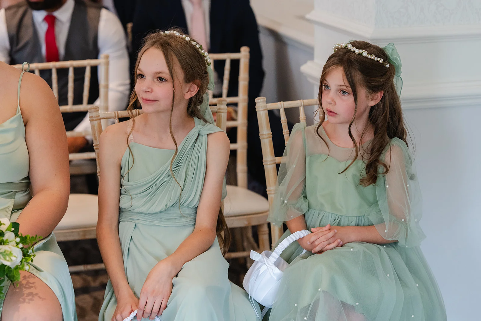 Two young bridesmaids wearing sage green dresses and matching pearl headbands sit attentively during the wedding ceremony.
