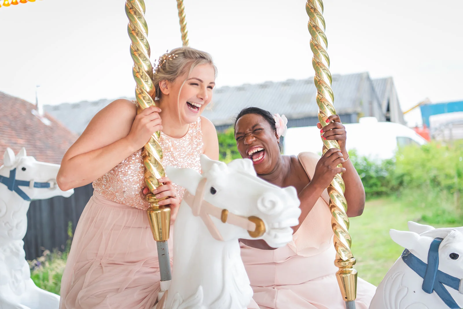 Two bridesmaids in pink dresses laugh joyfully while riding a vintage carousel at an outdoor wedding reception.