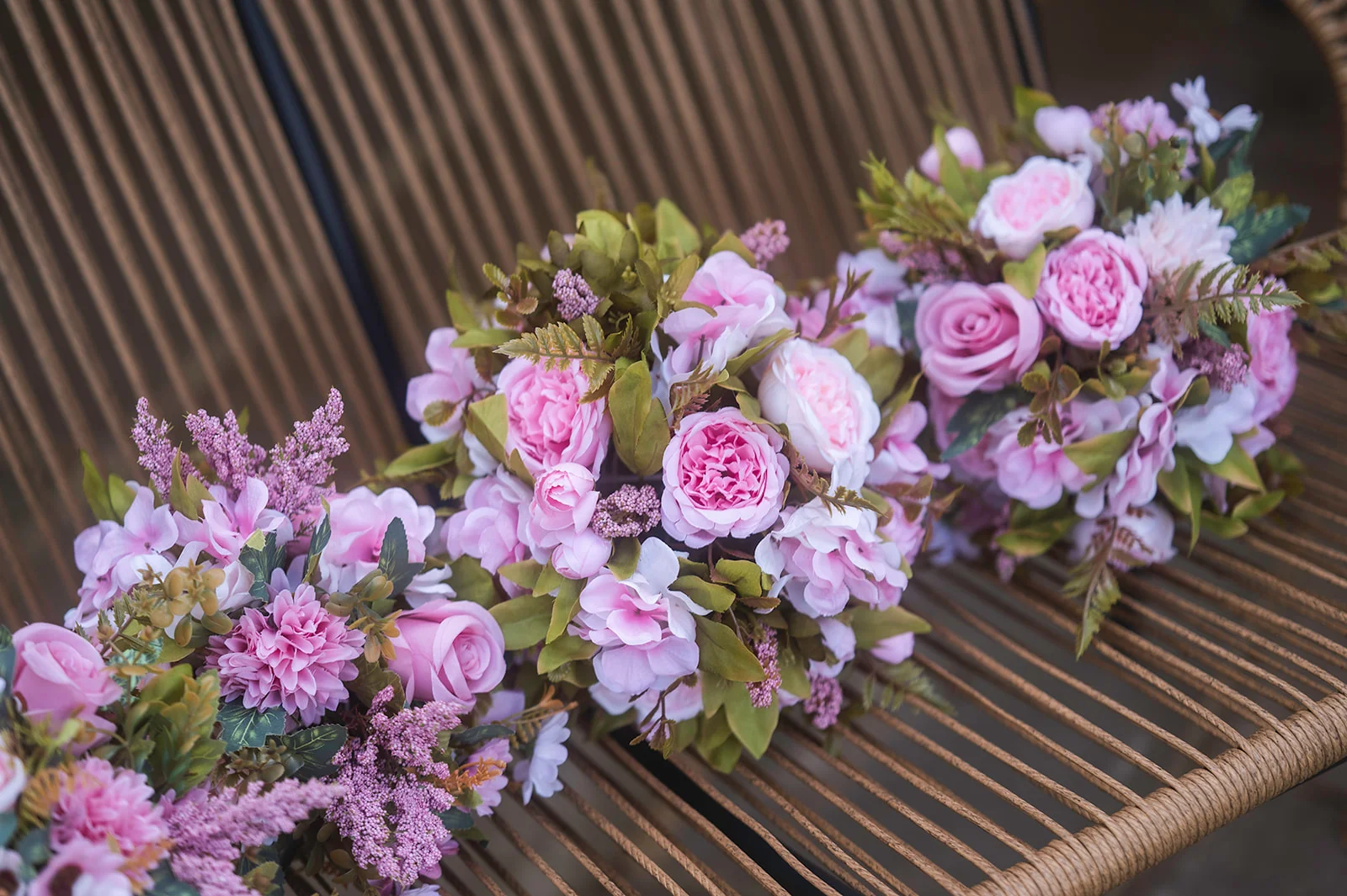Three matching hand-tied pink bridesmaid bouquets featuring garden roses and delicate greenery are arranged in a row on a woven rattan bench.