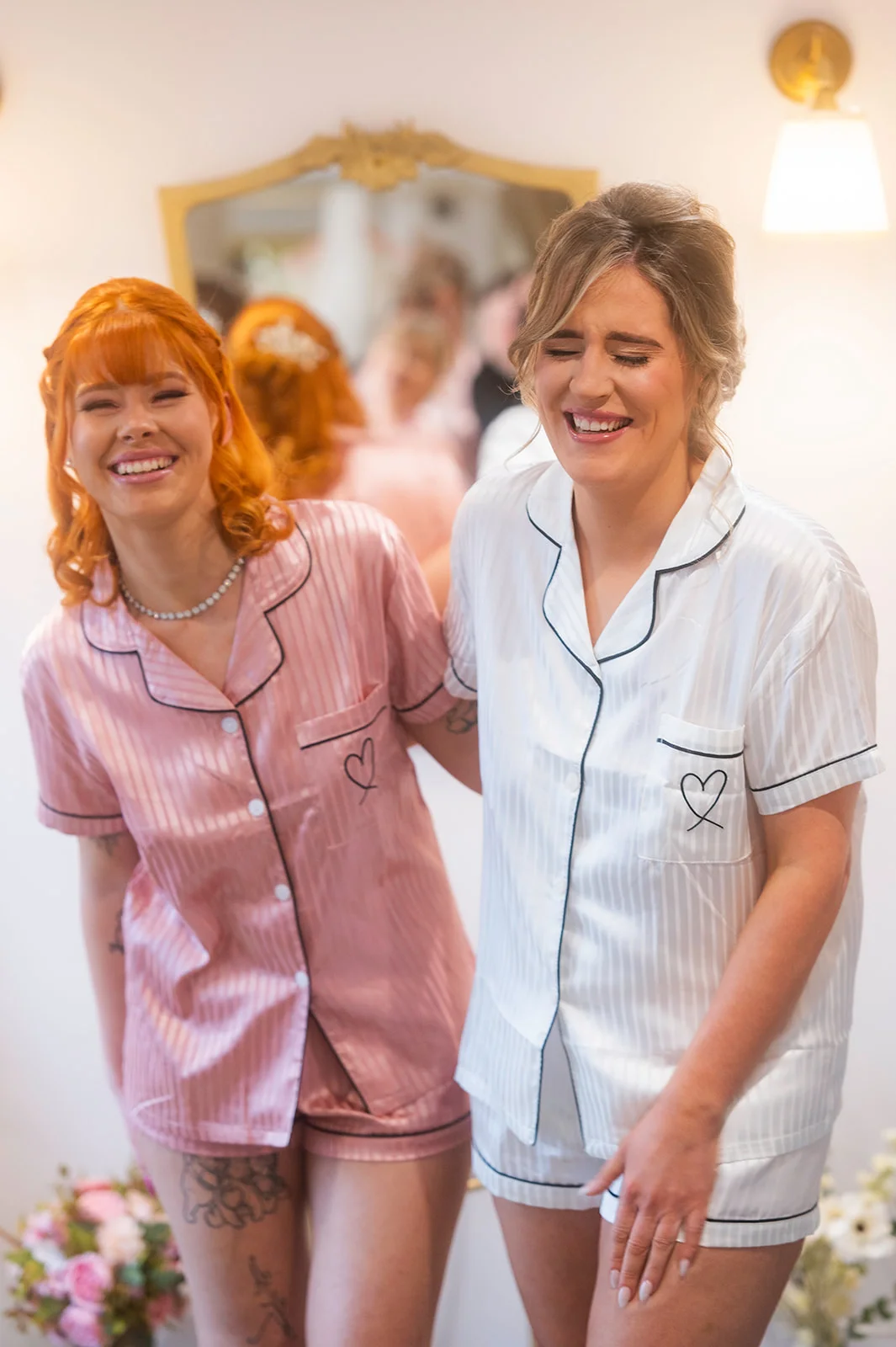 A bride in a white satin pyjama set and her bridesmaid in matching pink pyjamas share a joyful, laughing moment together indoors during morning bridal preparations.