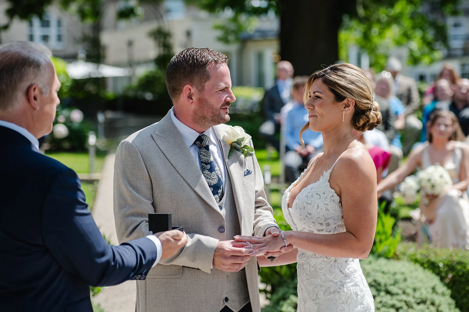 The groom places a ring on the bride's finger during a sunlit outdoor garden ceremony.